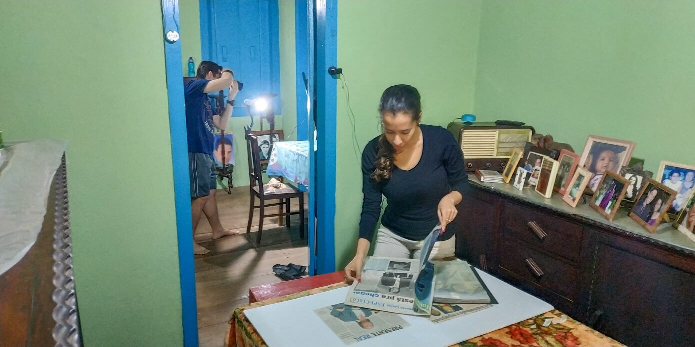 Luan Volpato fotografando recortes de jornal enquanto Sara Moreira folheia jornais para encontrar imagens e notícias de Roberto Carlos para serem fotografadas.