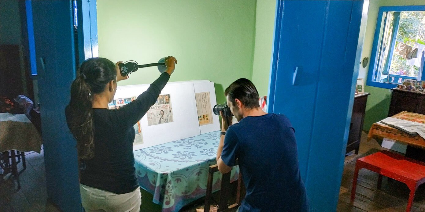 Luan Volpato fotografando recortes de jornal enquanto Sara Moreira aponta um abajur para iluminá-los.