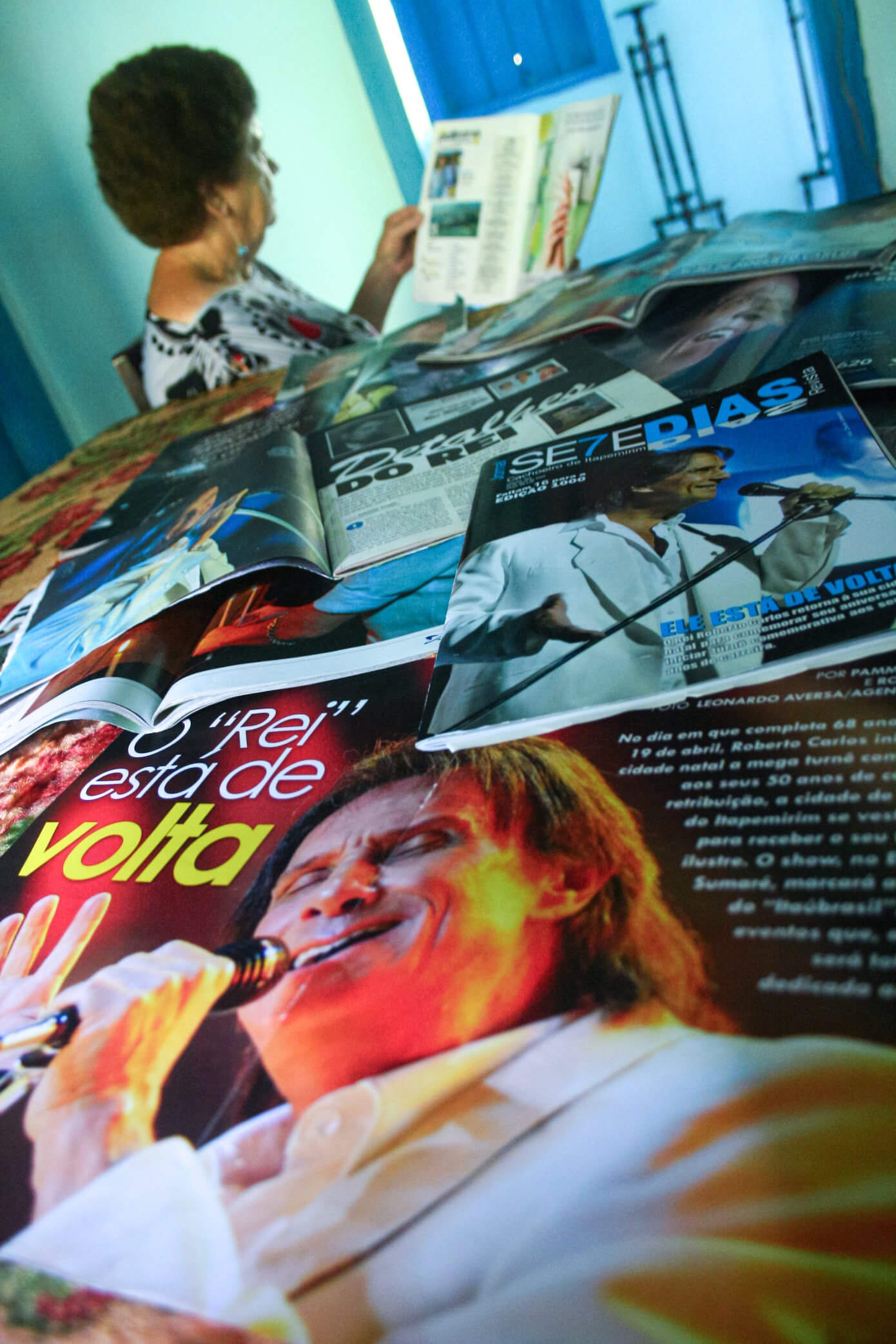 A imagem mostra Gercy Volpato lendo uma revista em meio a várias revistas espalhadas em uma mesa.