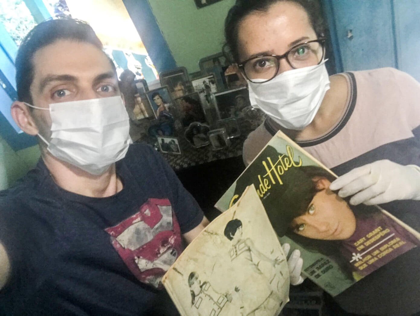 Luan Volpato e Sara Moreira, usando máscaras e luvas, seguram revistas da década de 50 do acervo de Gercy Volpato.